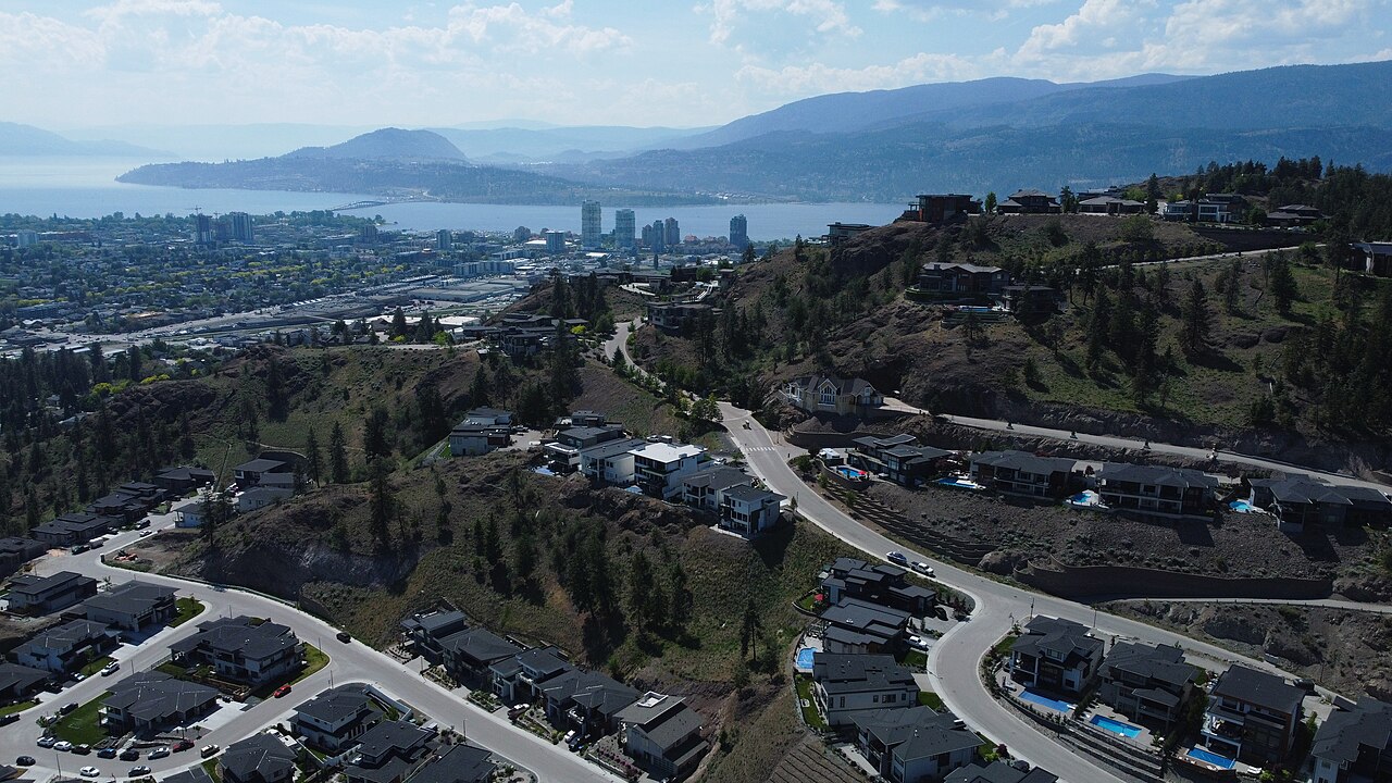 An aerial view of a city with buildings and roads on hillsides, with a large lake and mountains in the background under a partly cloudy sky.