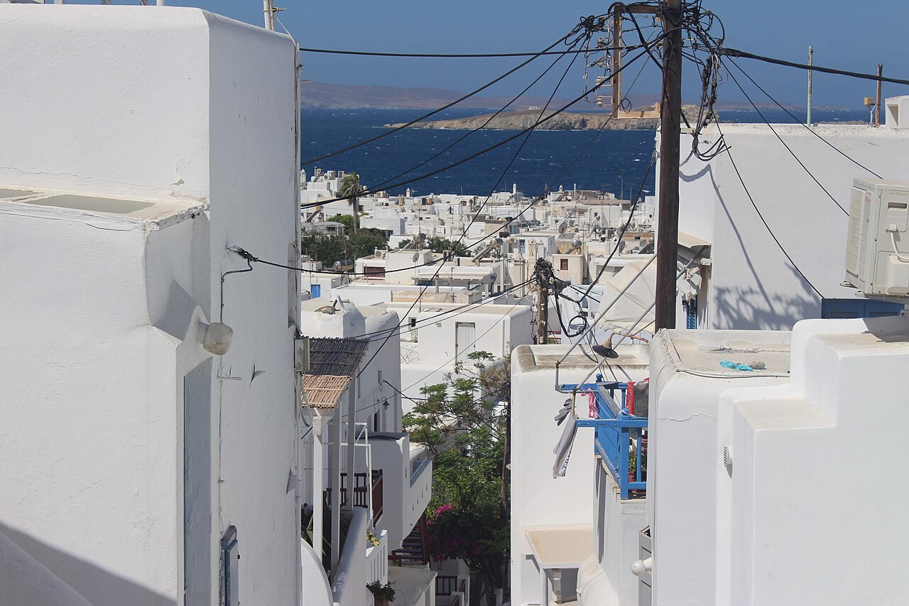 A narrow street winding down between white buildings, with many more white buildings and the sea in the distance.
