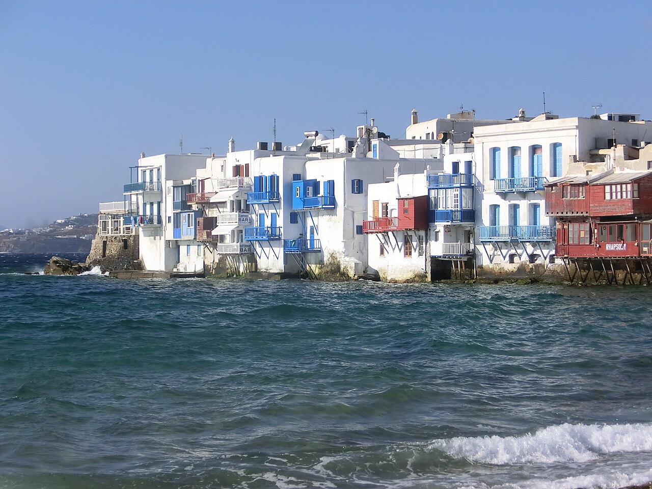 White buildings with blue and red balconies built directly on the water's edge, with waves crashing below.