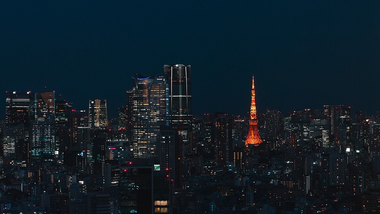 A dark night cityscape of lit office and apartment towers, with a glowing orange lattice tower standing among them.
