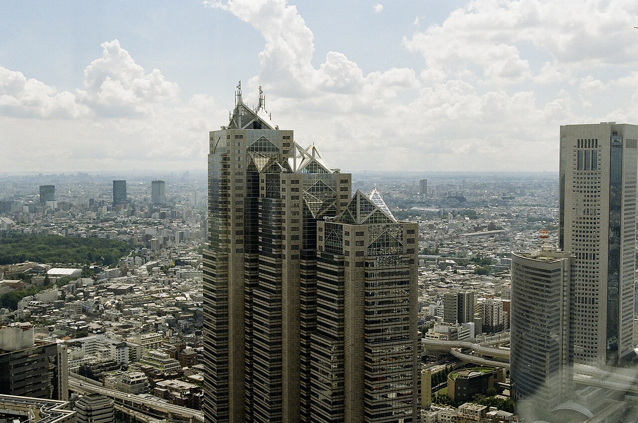 A three-stepped grey skyscraper with pyramid-shaped glass tops rising above a low, dense city that stretches to the horizon.