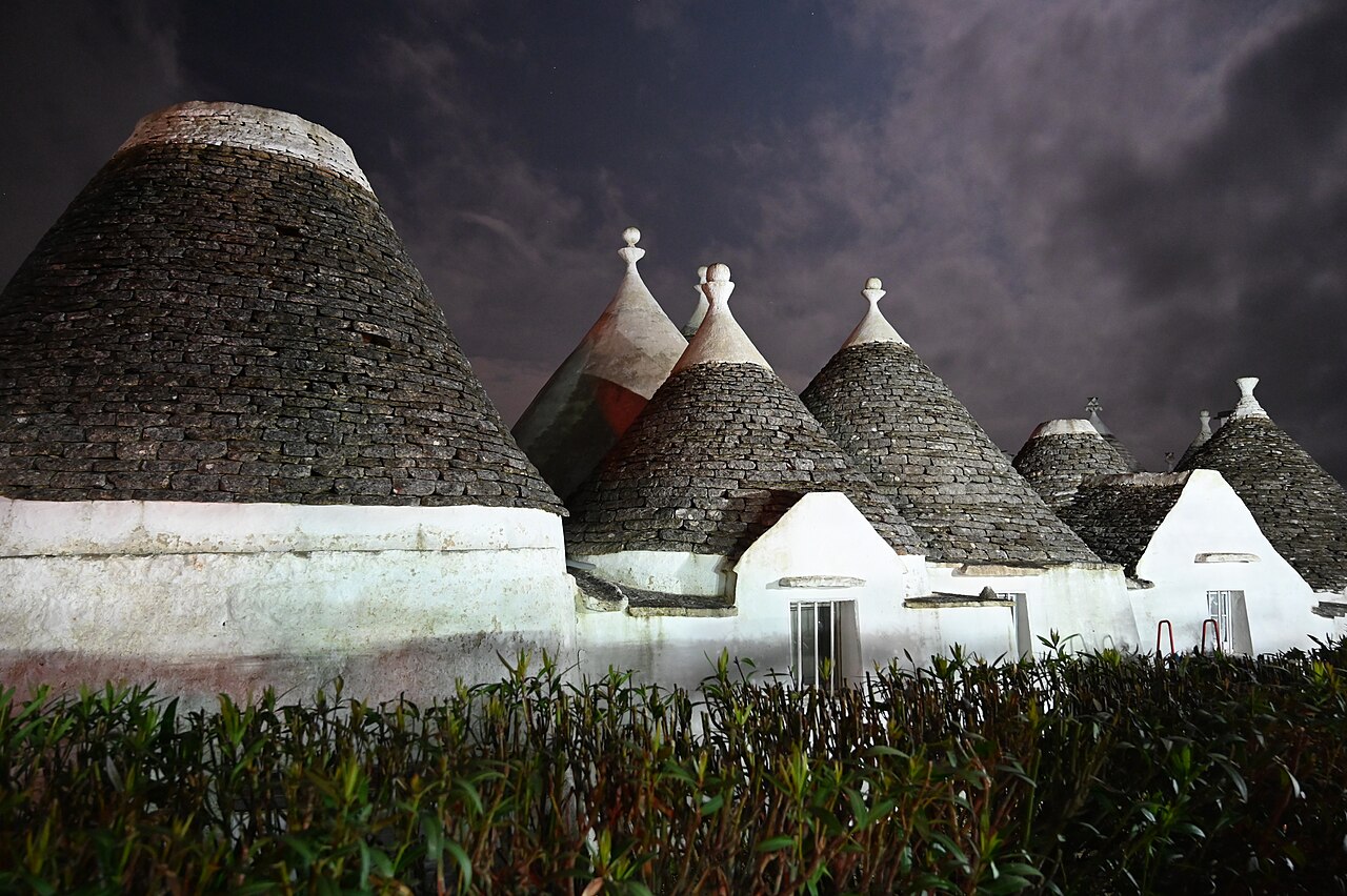 Several white, conical-roofed stone huts are visible behind a low hedge, under a dark, cloudy sky.