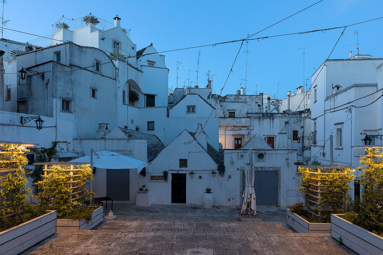 A small square with illuminated planters in the foreground, leading to a cluster of white buildings with various rooflines under a twilight sky.