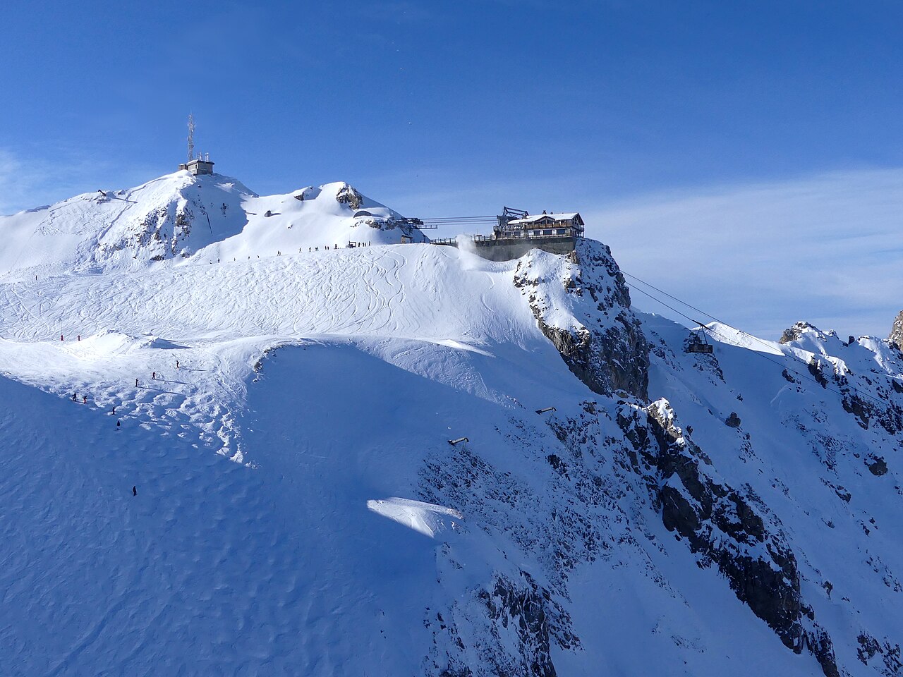 A snow-covered summit with a lift station on its ridge, above steep white slopes and a rocky face.