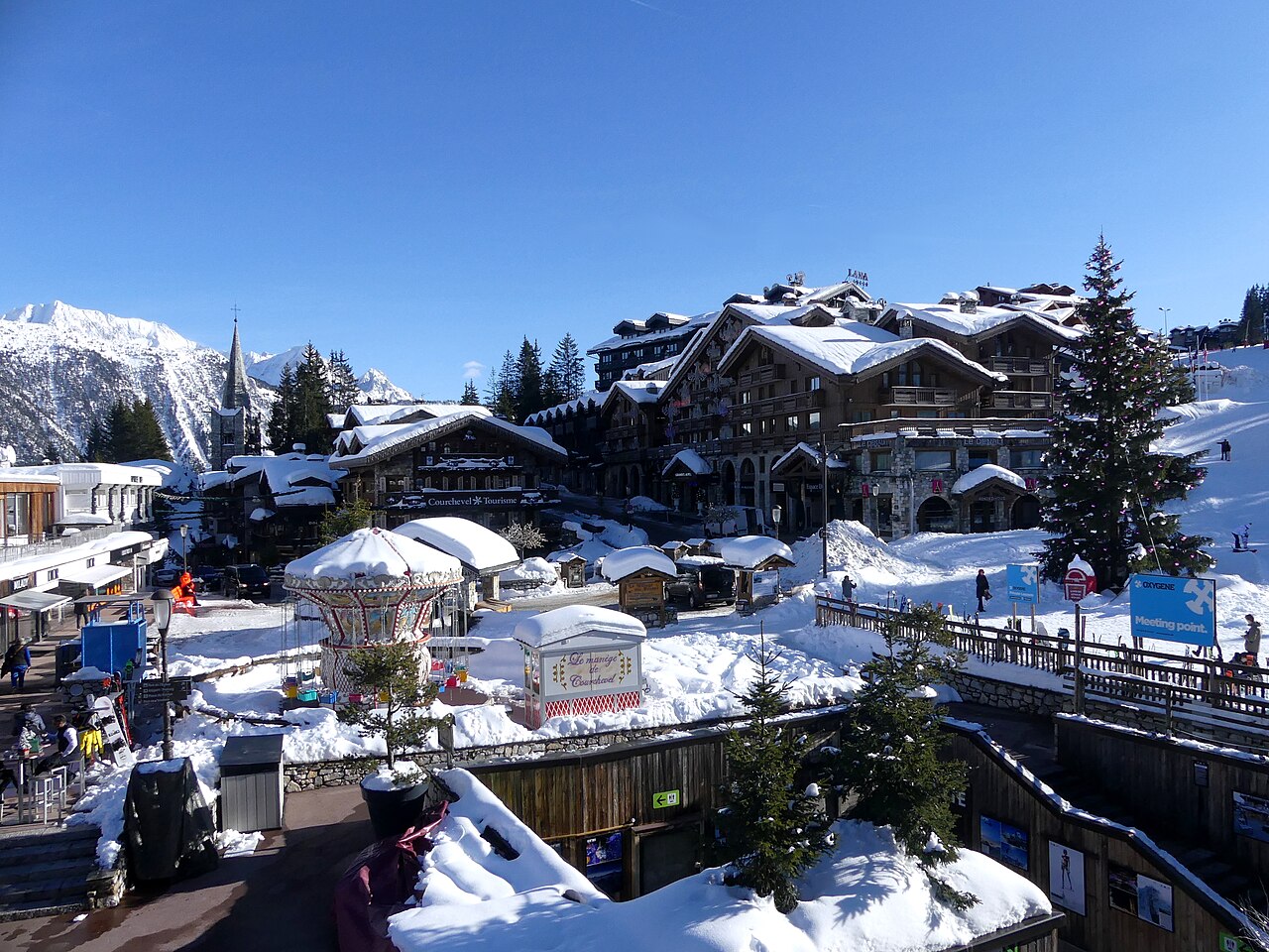 Large timber chalets with snow on their roofs above a square with small stalls and fir trees, under a deep blue sky.