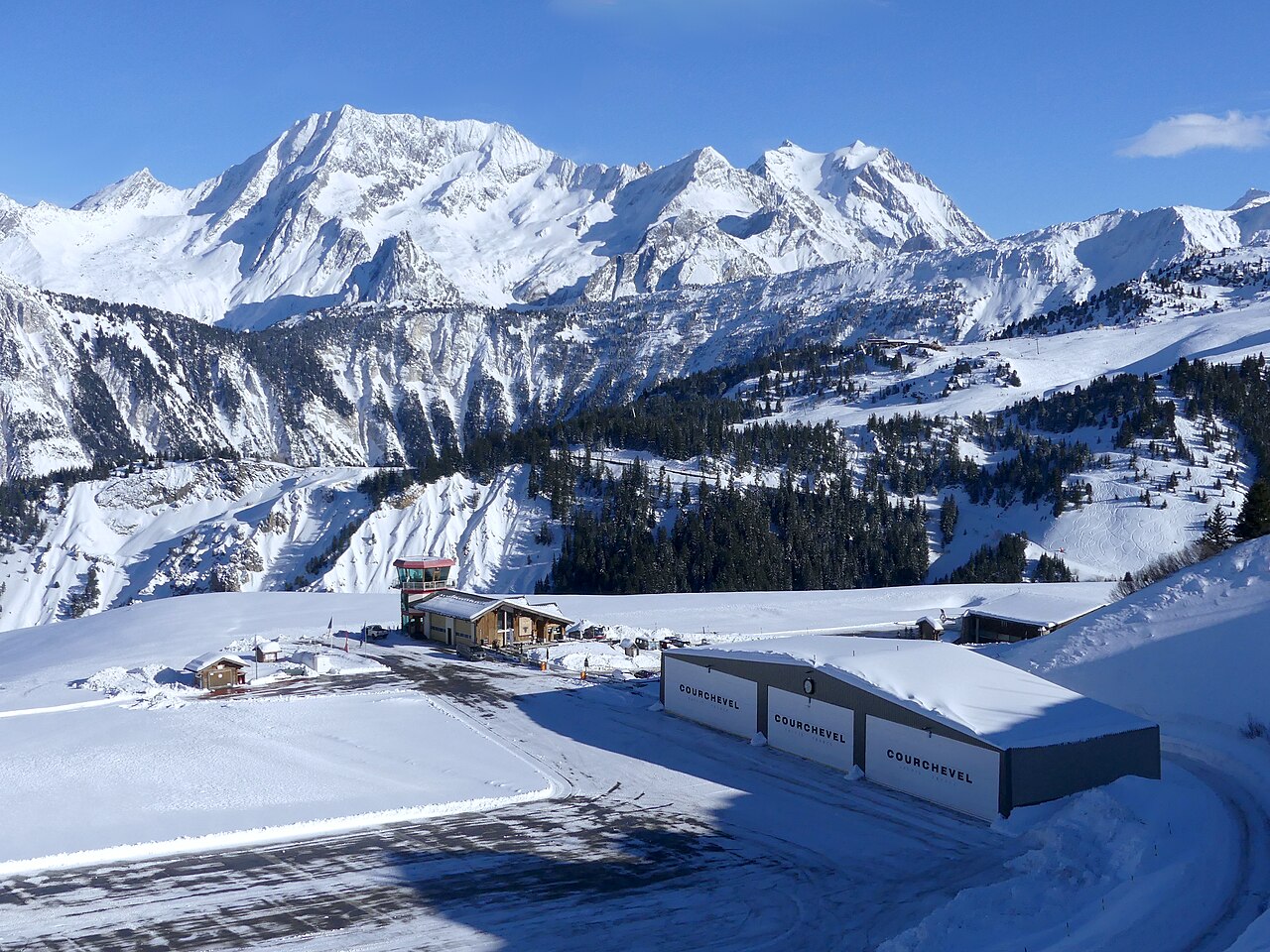 Three low hangars beside a cleared tarmac in the snow, with a wall of high white peaks behind.