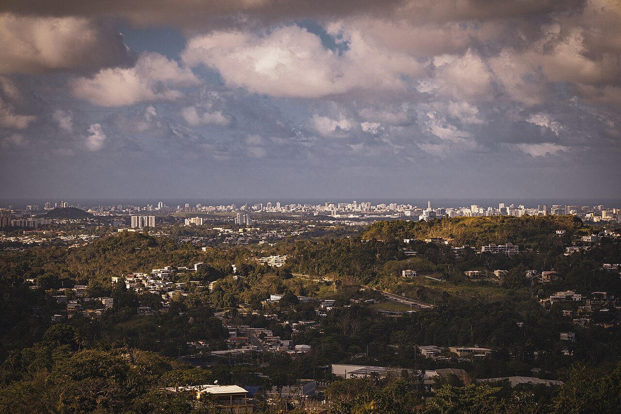 A wide shot of a city skyline in the distance, with rolling green hills and scattered buildings in the foreground under a cloudy sky.