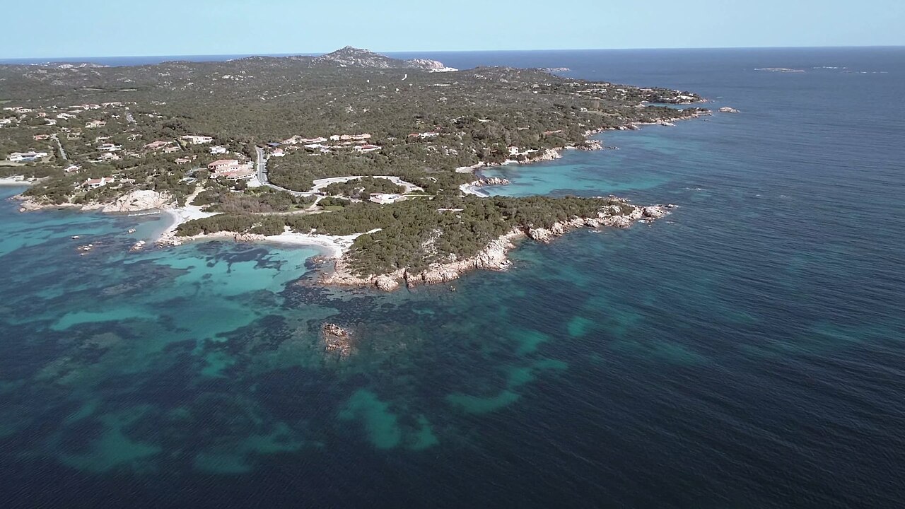 A rocky, scrub-covered headland with small coves, surrounded by clear water shading from turquoise to deep blue.