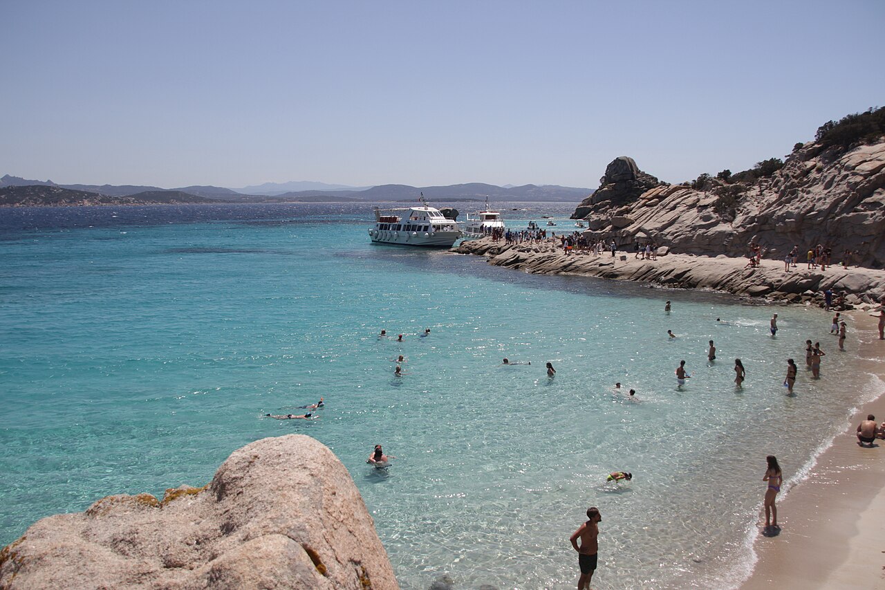 Clear turquoise shallows and pale sand between rounded granite rocks, with swimmers in the water and a boat near the shore.