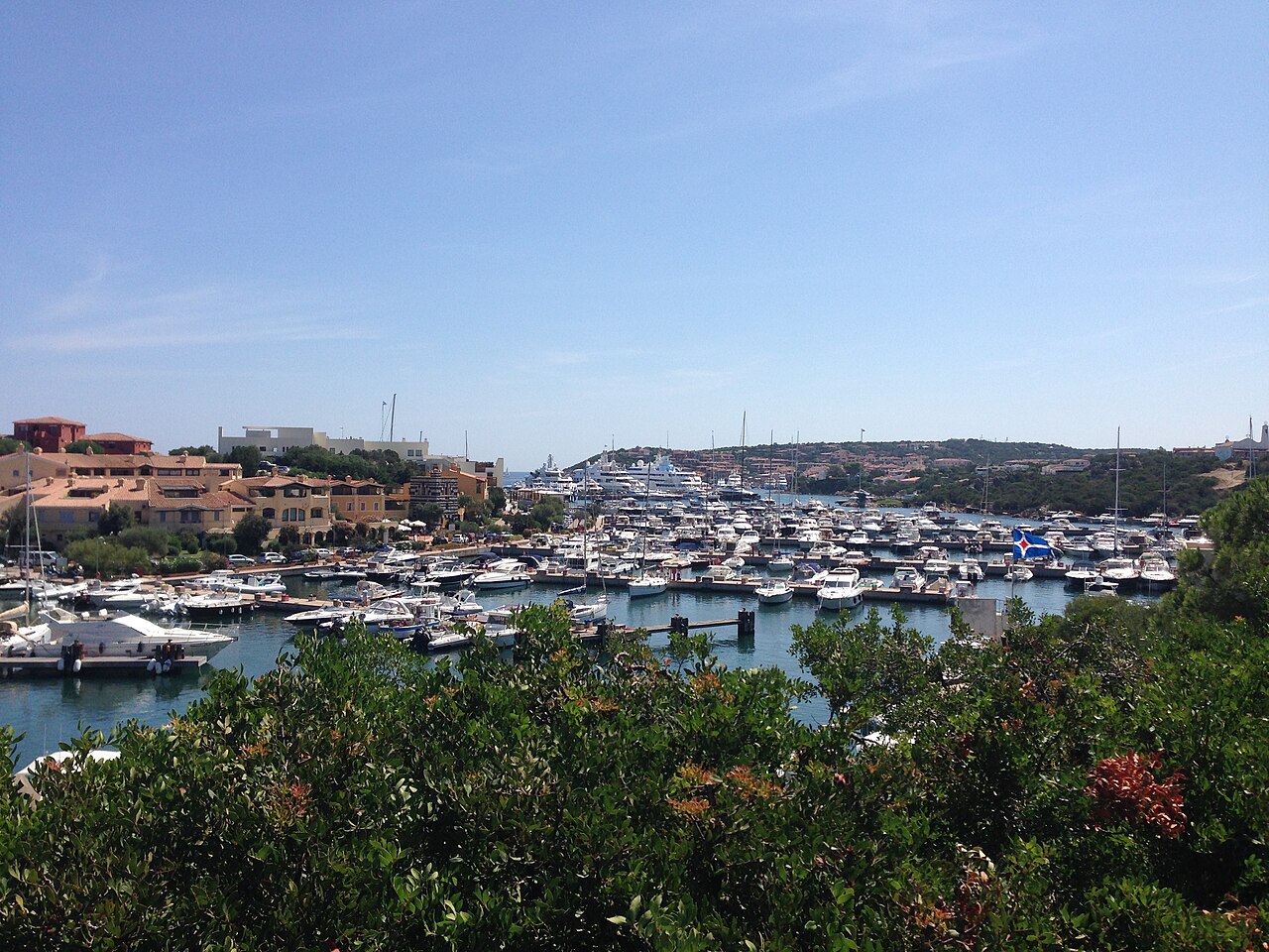 A sheltered bay filled with moored boats, with low terracotta-coloured buildings on the far shore and flowering shrubs in front.