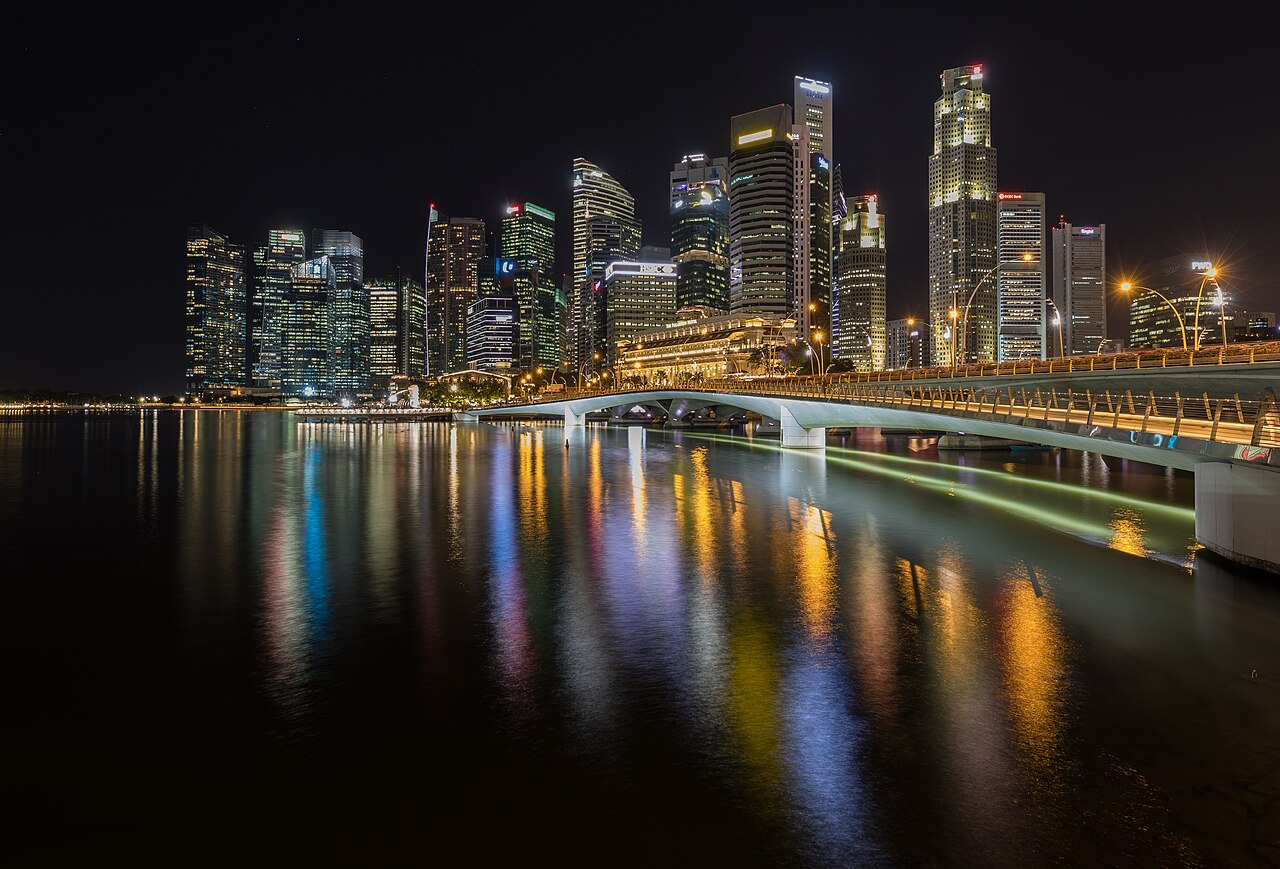 A night view of a city skyline with illuminated skyscrapers and a bridge over water, reflecting colorful lights.