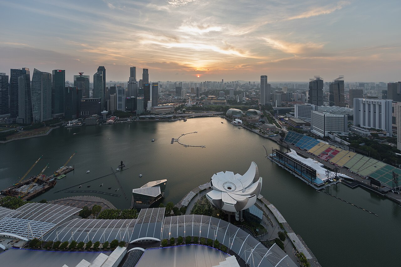 An aerial view of a city skyline at sunset, with skyscrapers surrounding a body of water under an orange sky.